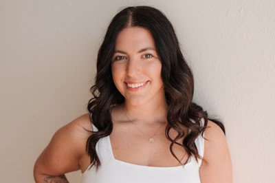 Maddie is standing in a white tank top and jeans, smiling with her hand on her hip