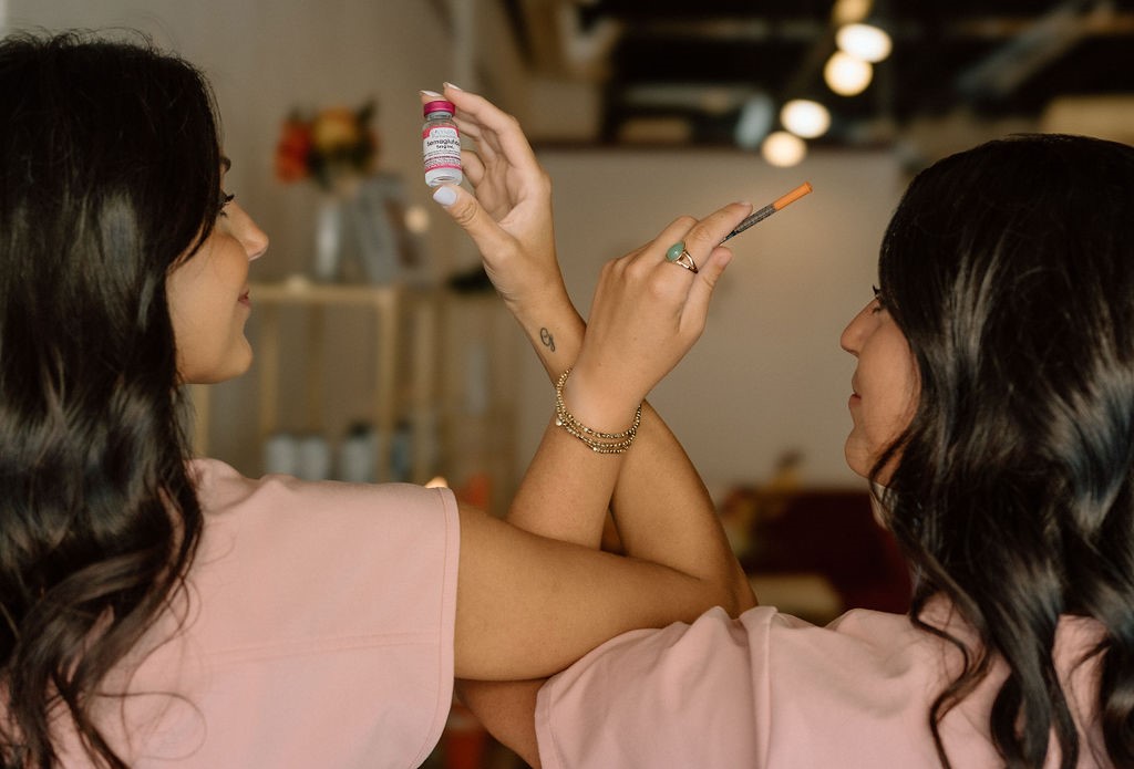 two woman, backs turned and one holding liquid med bottle and one holding a syringe