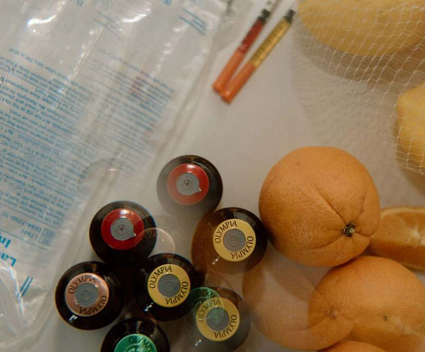 An overhead arrangement showing several brown supplement vials with colored caps, two syringes, a clear IV fluid bag, and a mesh bag of oranges with one sliced open. All items are placed on a light-colored surface, creating a clinical yet natural wellness theme