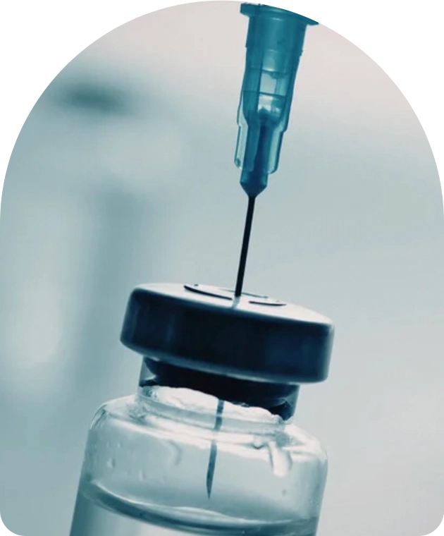 A close-up view of a syringe needle inserted through the rubber stopper of a clear medical vial, drawing up liquid. The background is softly blurred in cool blue tones.
