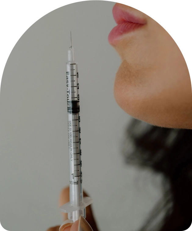 A close-up view of a hand holding a thin medical syringe upright. Only the lower half of a person’s face is visible near the needle, with the background softly blurred.