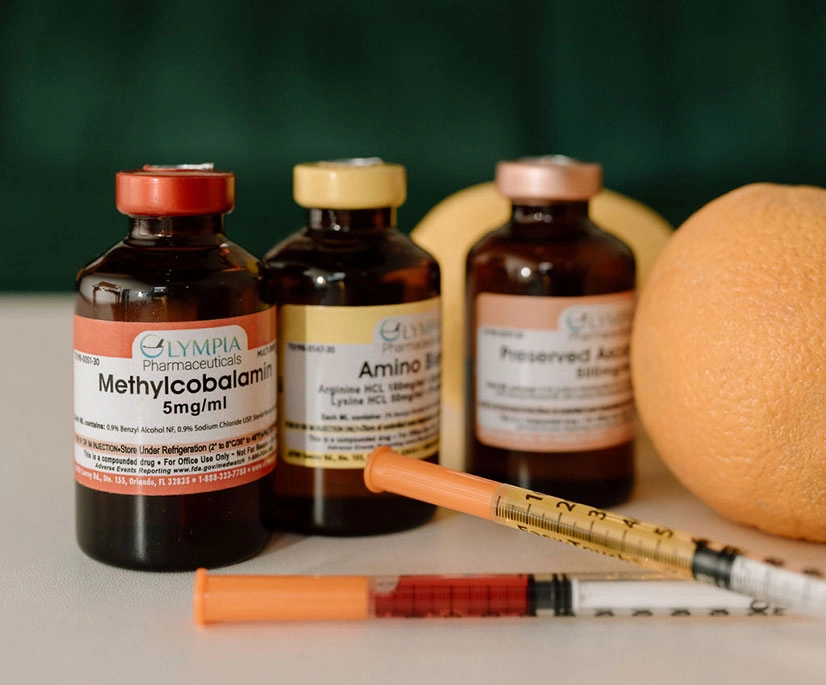 Three brown glass vials labeled ‘Methylcobalamin,’ ‘Amino Blend,’ and ‘Preserved Aqueous’ sit on a tabletop beside whole oranges. Two syringes lie in front of the vials, and a blurred dark green background gives the scene a clean, professional wellness setting.