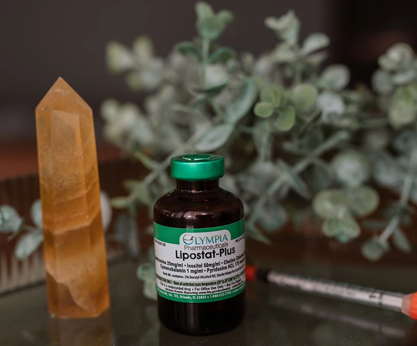 A brown glass vial labeled ‘Olympia Pharmaceuticals Lipostat‑Plus’ with a green cap sits on a reflective surface next to a tall orange crystal point. A syringe lies partially visible to the right, and soft green eucalyptus leaves fill the background.